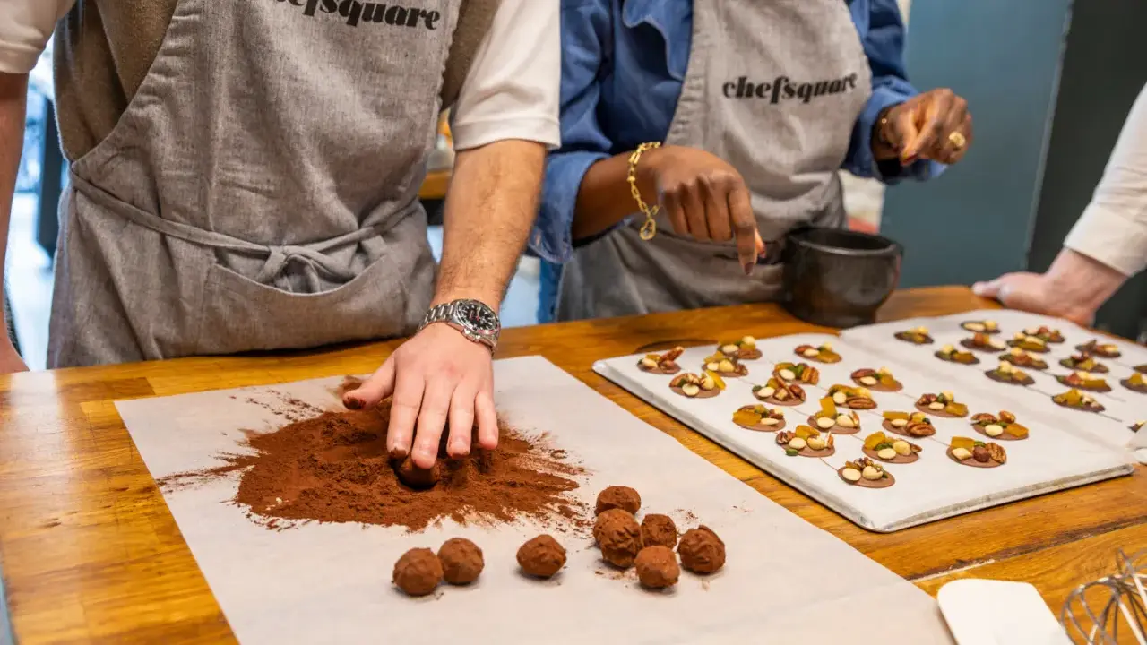 Des mains préparent du chocolat fait maison, roulant des truffes dans du cacao. Sur une plaque, des chocolats décorés de fruits secs attendent.