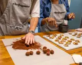 Des mains préparent du chocolat fait maison, roulant des truffes dans du cacao. Sur une plaque, des chocolats décorés de fruits secs attendent.