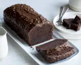 Un généreux gâteau au chocolat haut, nappé d'un glaçage croquant et parsemé de noisettes. Deux tranches sont déjà coupées, prêtes à être dégustées.