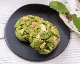Deux cookies clairs au matcha, parsemés de morceaux de chocolat blanc, sur une assiette noire, à côté d'une tasse de thé et de feuilles de menthe.