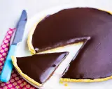Une part de la meilleure recette de tarte au chocolat, avec une ganache brillante sur une pâte dorée. Un couteau bleu est à côté.