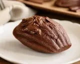 Un biscuit madeleine au chocolat, nappé de glaçage et parsemé de copeaux de chocolat, sur une assiette blanche.