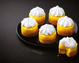 Six mini tarte au citron, topped with fluffy meringue, sit on a black plate. One has a bite taken out.