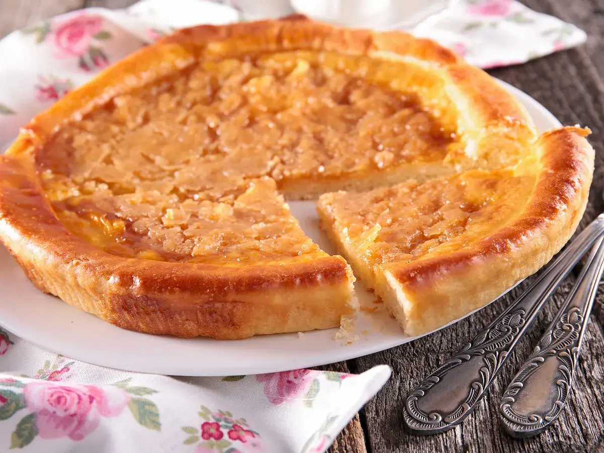 Une délicieuse tarte à la cassonade, dorée et appétissante, est servie sur une assiette blanche, prête à être dégustée avec des couverts ornés.