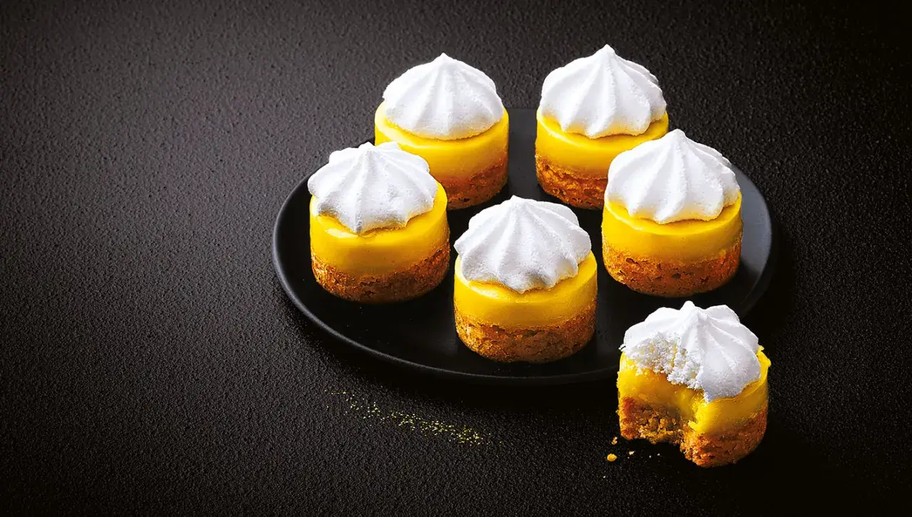Six mini tarte au citron, topped with fluffy meringue, sit on a black plate. One has a bite taken out.