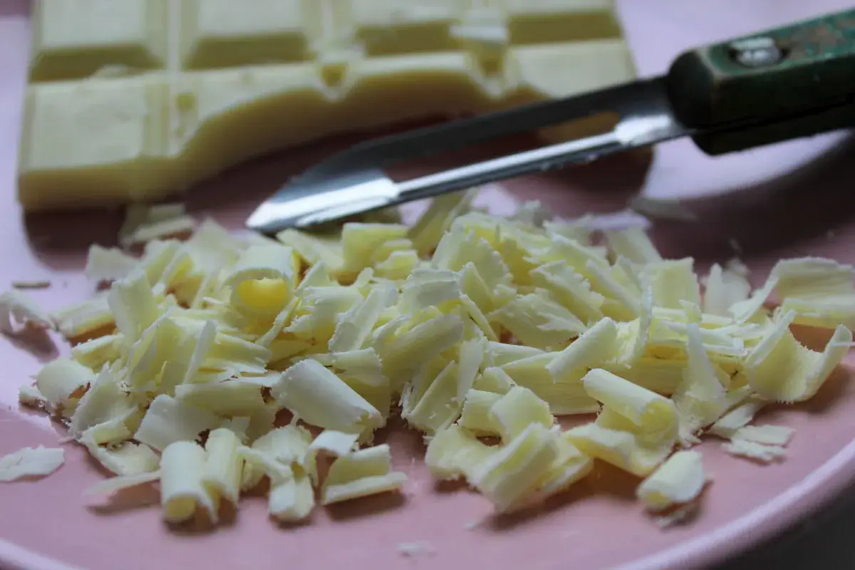 Comment râper du chocolat blanc : une barre de chocolat et des copeaux sur une assiette rose, avec un économe.