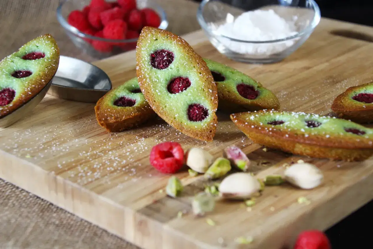 Financiers pistache framboise, saupoudrés de sucre glace, avec des framboises et des pistaches éparpillées sur une planche en bois.