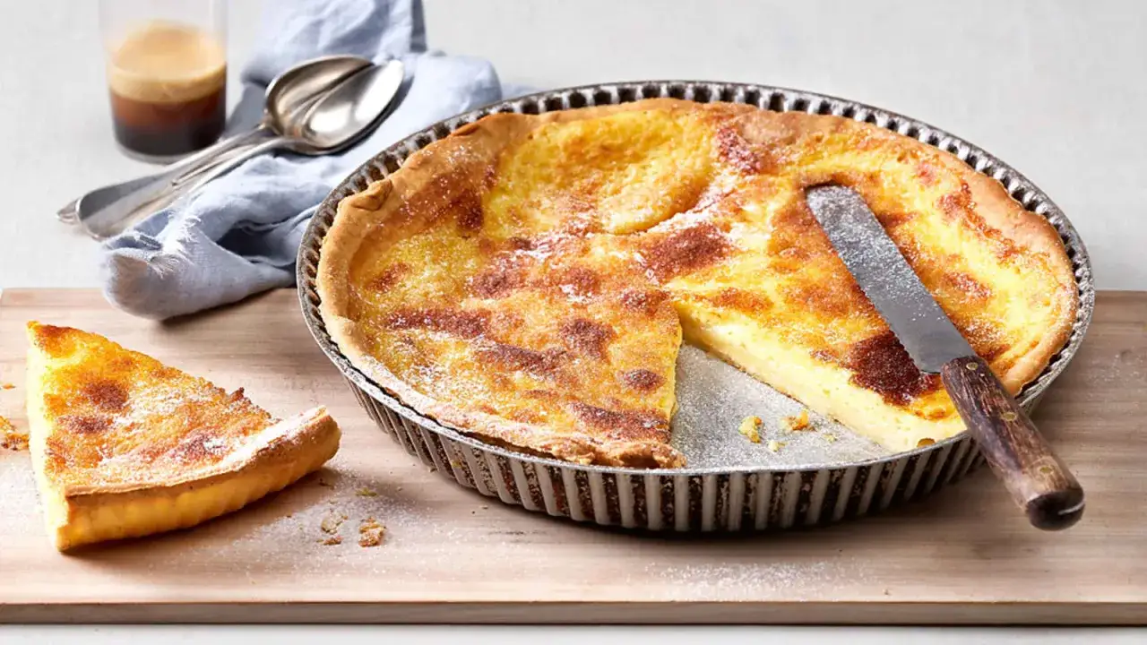 Une part de tarte à la crème de grand-mère, dorée et saupoudrée de sucre glace, repose sur une planche en bois. Une autre part est encore dans le moule.