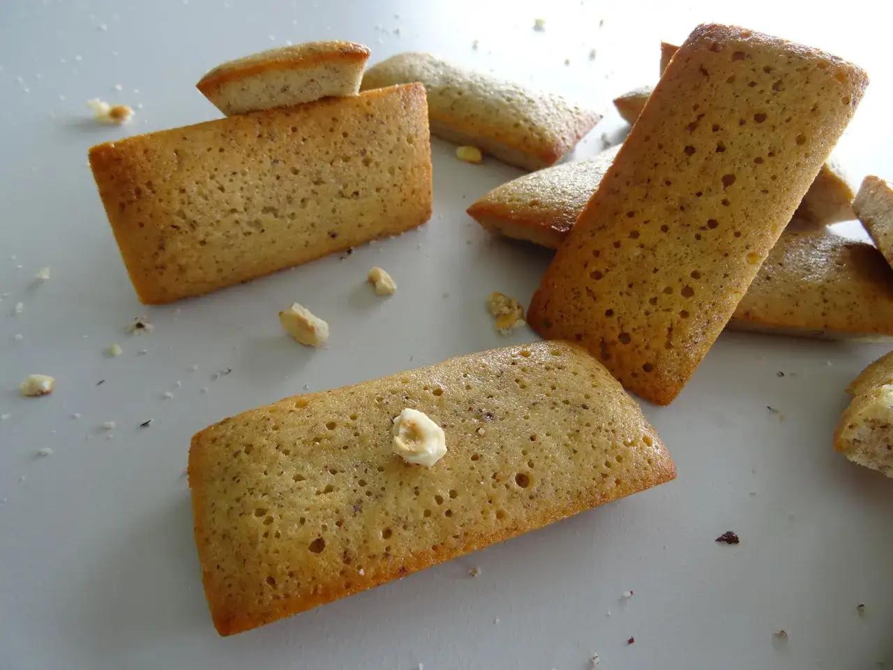 Plusieurs financiers aux noisettes dorés, parsemés de miettes et de quelques noisettes entières, sur fond clair.