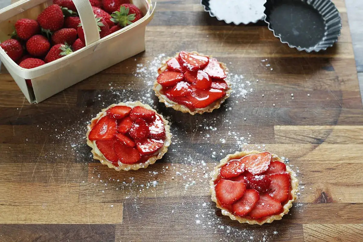 Trois mini tartelettes fraise, garnies de fruits rouges frais et de crème pâtissière, saupoudrées de sucre glace sur une planche en bois.