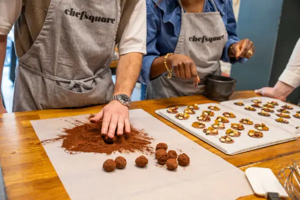 Des mains préparent du chocolat fait maison, roulant des truffes dans du cacao. Sur une plaque, des chocolats décorés de fruits secs attendent.