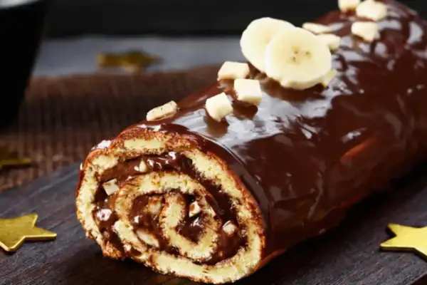 Bûche roulée au chocolat, garnie de crème et recouverte d'un glaçage chocolat beurre brillant, décorée de rondelles de banane et de noisettes concassées.