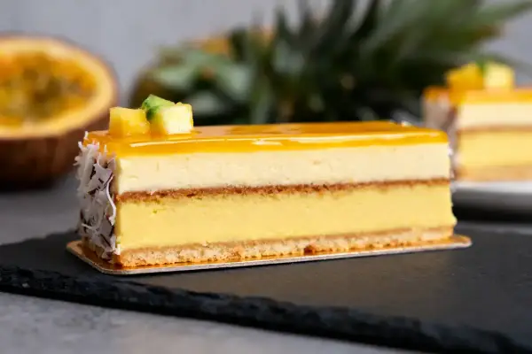 Un délice tropical : un entremets aux couches crémeuses, surmonté d'un glacage miroir jaune éclatant, décoré de fruits et de noix de coco râpées.