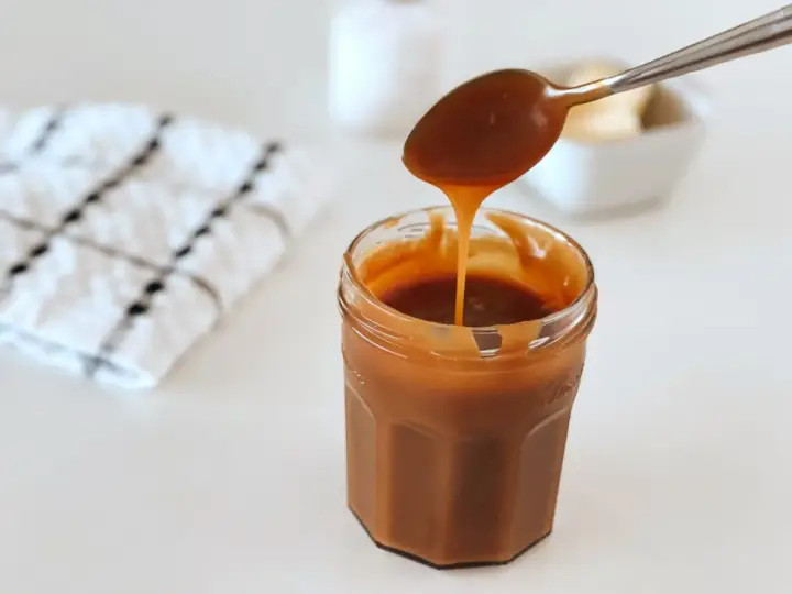 Une cuillère verse du caramel beurre salé dans un pot. Le beurre doux est visible en arrière-plan.