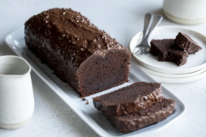 Un généreux gâteau au chocolat haut, nappé d'un glaçage croquant et parsemé de noisettes. Deux tranches sont déjà coupées, prêtes à être dégustées.