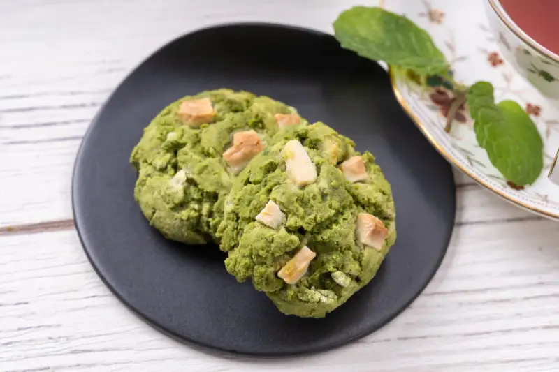 Deux cookies clairs au matcha, parsemés de morceaux de chocolat blanc, sur une assiette noire, à côté d'une tasse de thé et de feuilles de menthe.